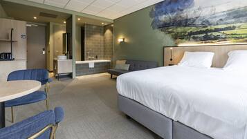Een hotelkamer met een groot bed, een kleine bank, een bureau met een stoel, een badkamer met douche en een schilderij aan de muur.
