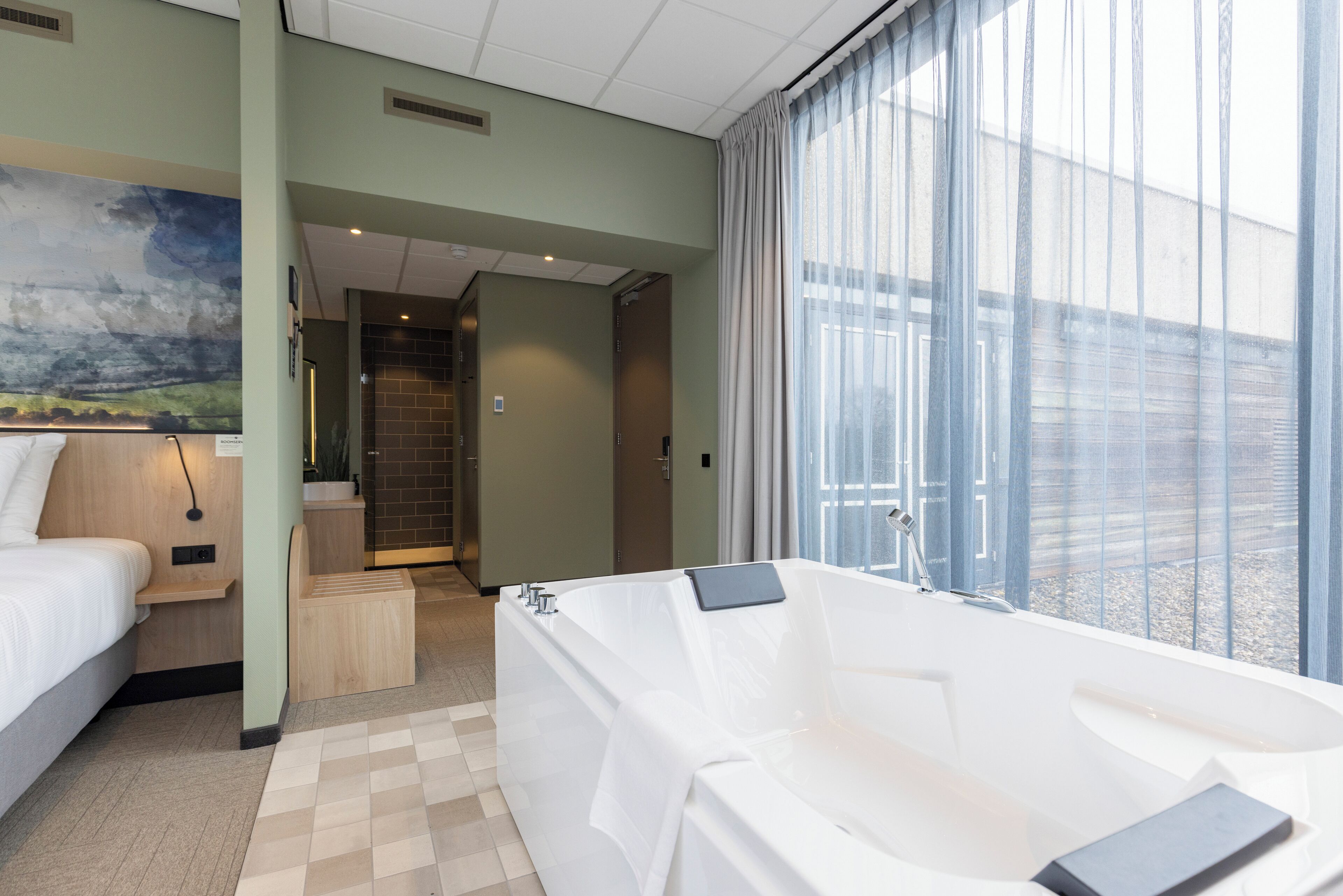 A modern hotel room with a large bathtub, a bed, and a view of the city.