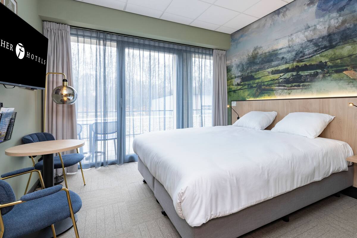 A hotel room with a large bed, a desk, and a chair. There is a painting on the wall and a window with curtains.