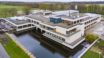 Un edificio moderno con terraza en la azotea, un cuerpo de agua en primer plano y un estacionamiento.