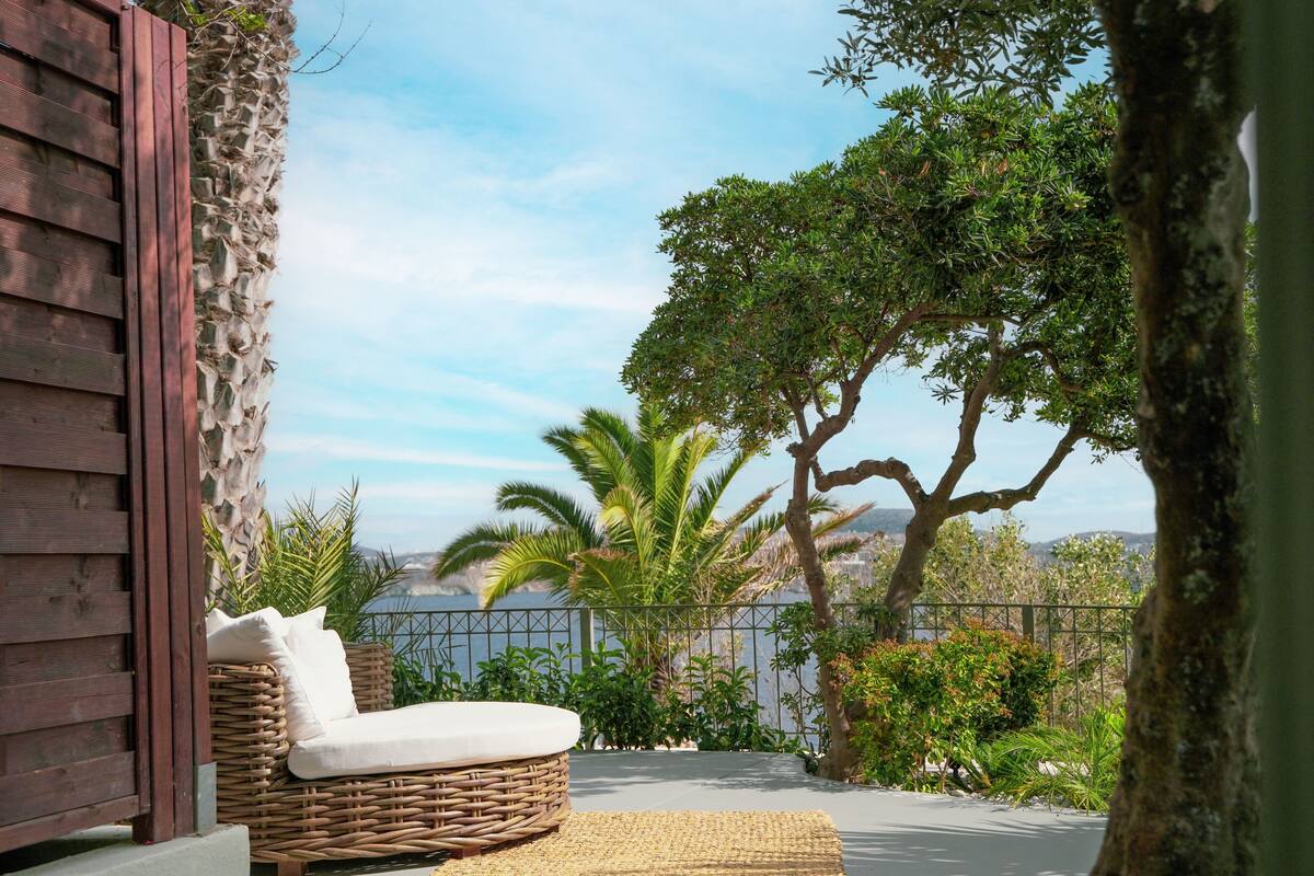 A wicker lounge chair with cushions on a balcony overlooking a body of water, surrounded by lush greenery and a stone wall.