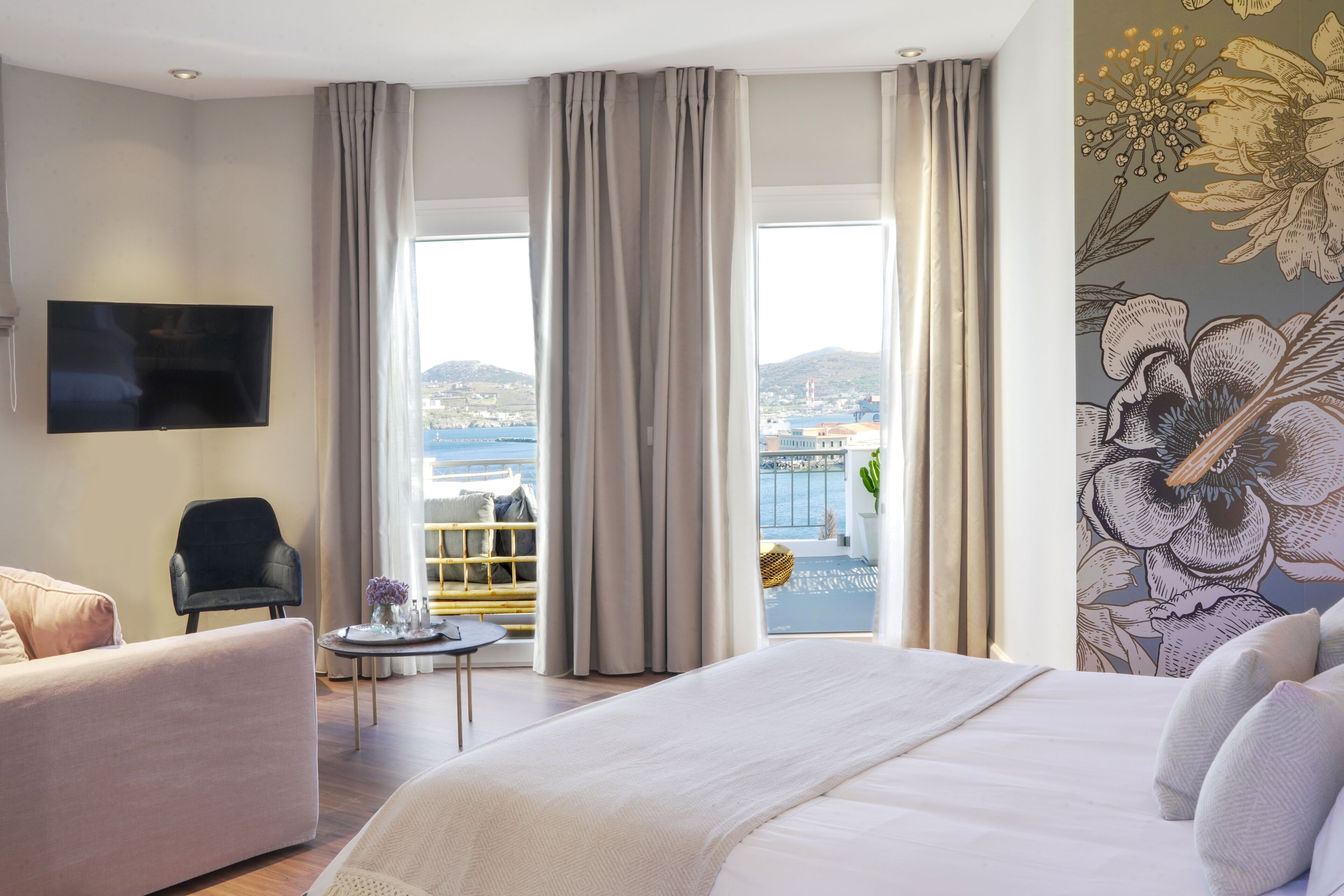 A hotel room with a large bed, a TV mounted on the wall, a chair, a small table, and a balcony with a view of the sea.