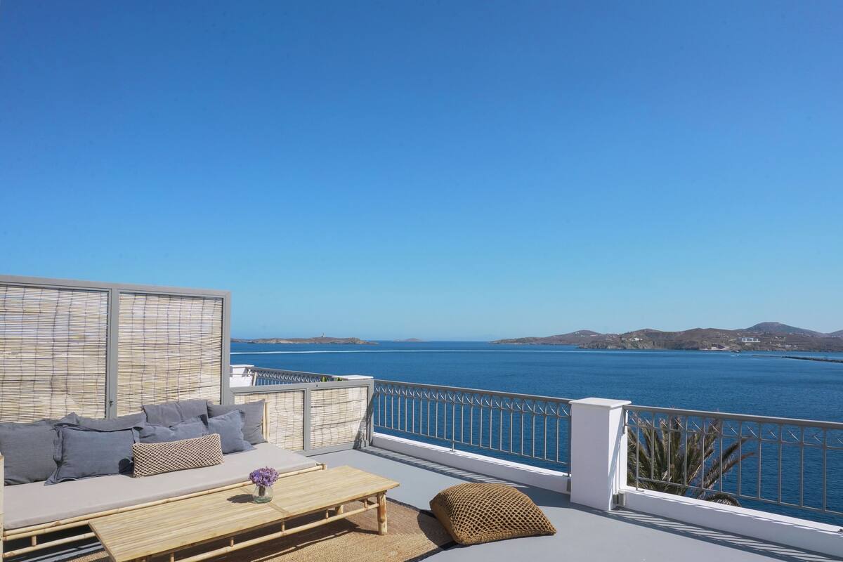A balcony with a sofa, cushions, a table, and a view of the sea and hills.
