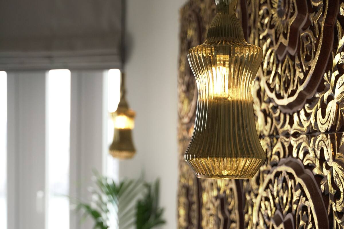 A bed with white bedding and decorative pillows, a wall with intricate golden carvings, and a hanging light fixture.