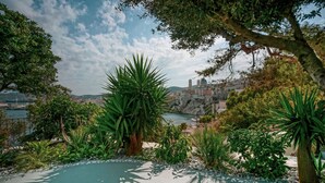 A landscaped garden with a pool, surrounded by various plants and trees, overlooking a coastal town and a body of water. - Pafsilipon Suites (Syros)