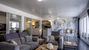 A modern living room with a grey sofa, a wooden coffee table, and a dining area with white chairs and a table. - Beachfront & Town Center • 2 Bedrooms • 2 Ensuite Baths (Le Diamant)
