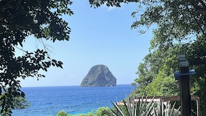 A view of a coastal area with a prominent rocky island in the distance, surrounded by lush greenery and a clear blue sea. - Beachfront & Town Center • 2 Bedrooms • 2 Ensuite Baths (Le Diamant)
