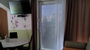 Una habitación con una mesa de comedor, sillas, un televisor fijado a la pared, un ventanal corredero con persianas y un sofá.