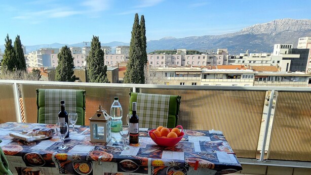 Un balcón con una mesa dispuesta para una comida, que incluye una botella de vino, copas y un bol de naranjas. El fondo muestra edificios y montañas.
