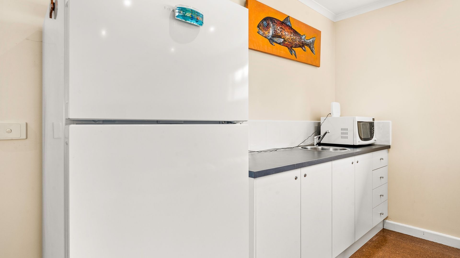 A kitchen with a white refrigerator, a microwave, and a sink. There is a painting of a fish on the wall.