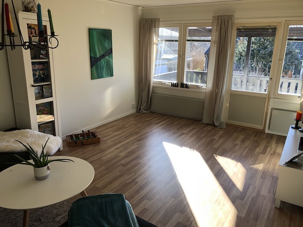 Une pièce spacieuse avec un plancher en bois, un canapé blanc, une petite table ronde agrémentée d’une plante, une bibliothèque et une grande fenêtre avec des rideaux.