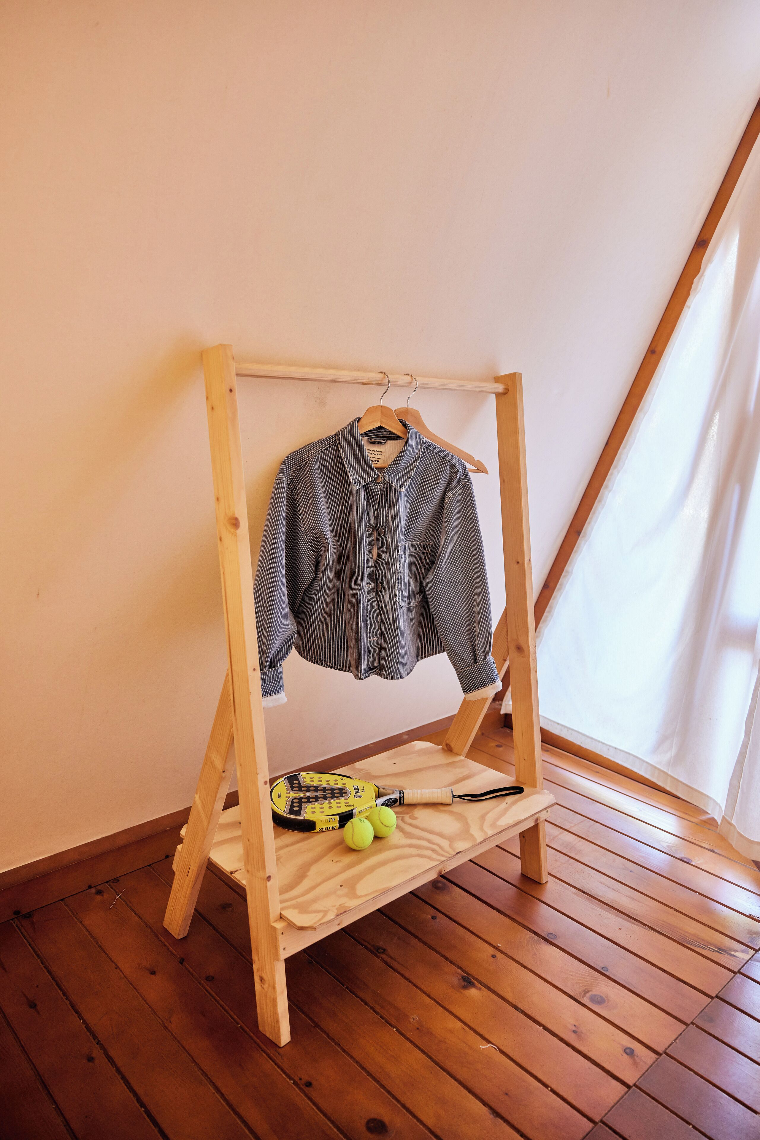 Un perchero de madera con una camisa a rayas, una raqueta de tenis y una pelota de tenis sobre un piso de madera en una habitación con techo inclinado.