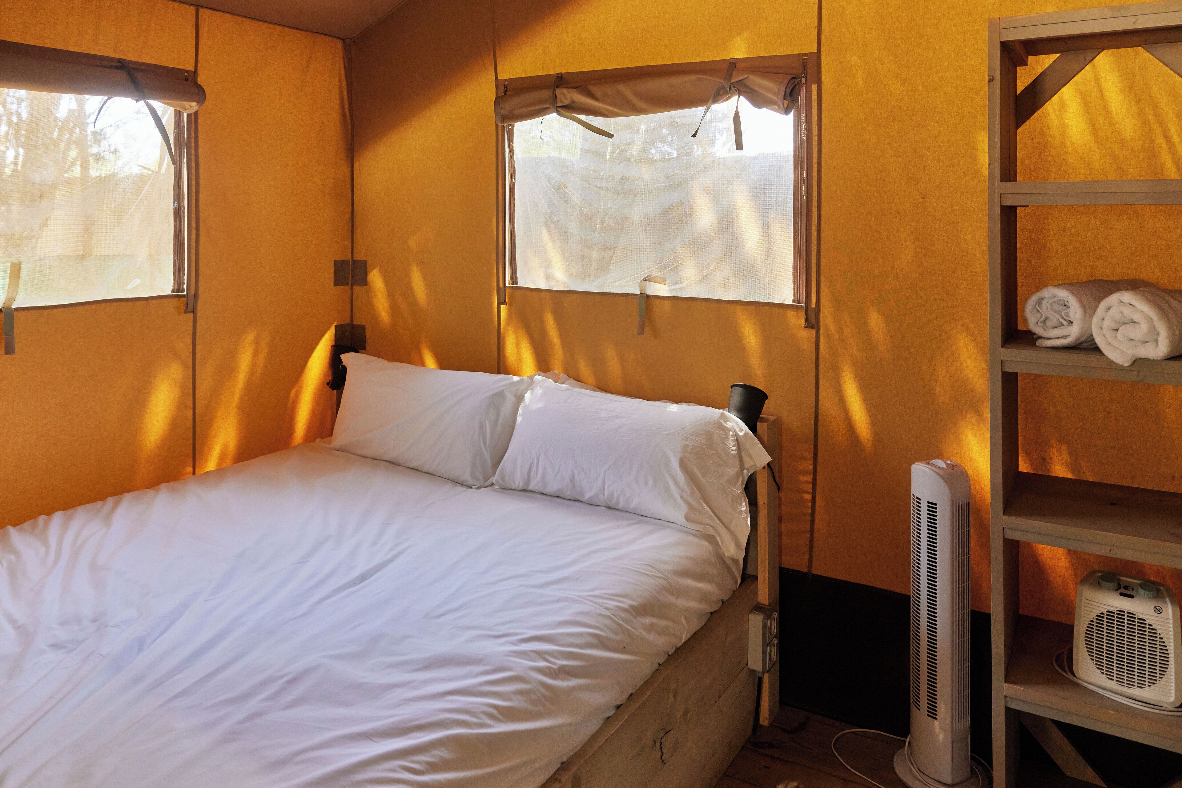 A single bed with white bedding, two windows with curtains, a wooden shelf with rolled towels, and a white portable heater.