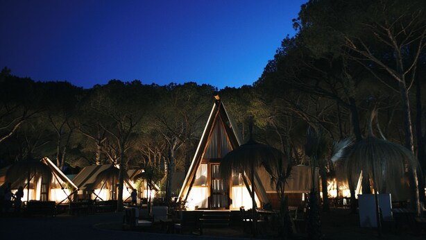 A series of wooden cabins with thatched roofs, surrounded by trees, at night. - TAIGA Conil (Conil de la Frontera)