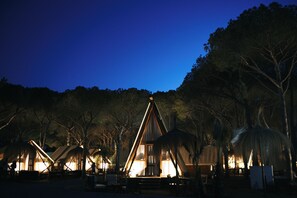 A series of wooden cabins with thatched roofs, surrounded by trees, at night. - TAIGA Conil (Conil de la Frontera)