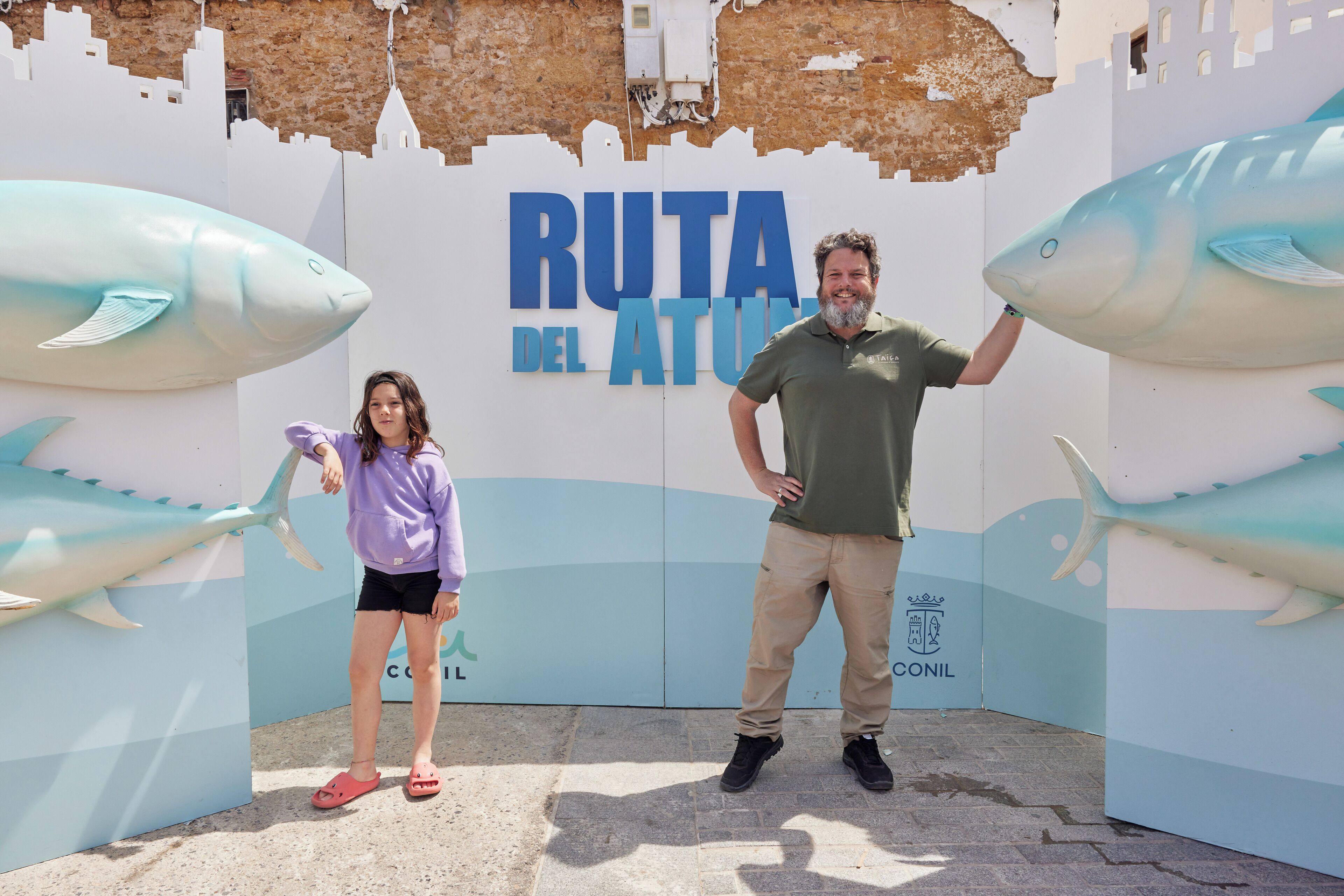 Ein Mann und ein Kind stehen vor einem Schild mit der Aufschrift "RUTA DEL ATUN" und großen Fischabbildungen.