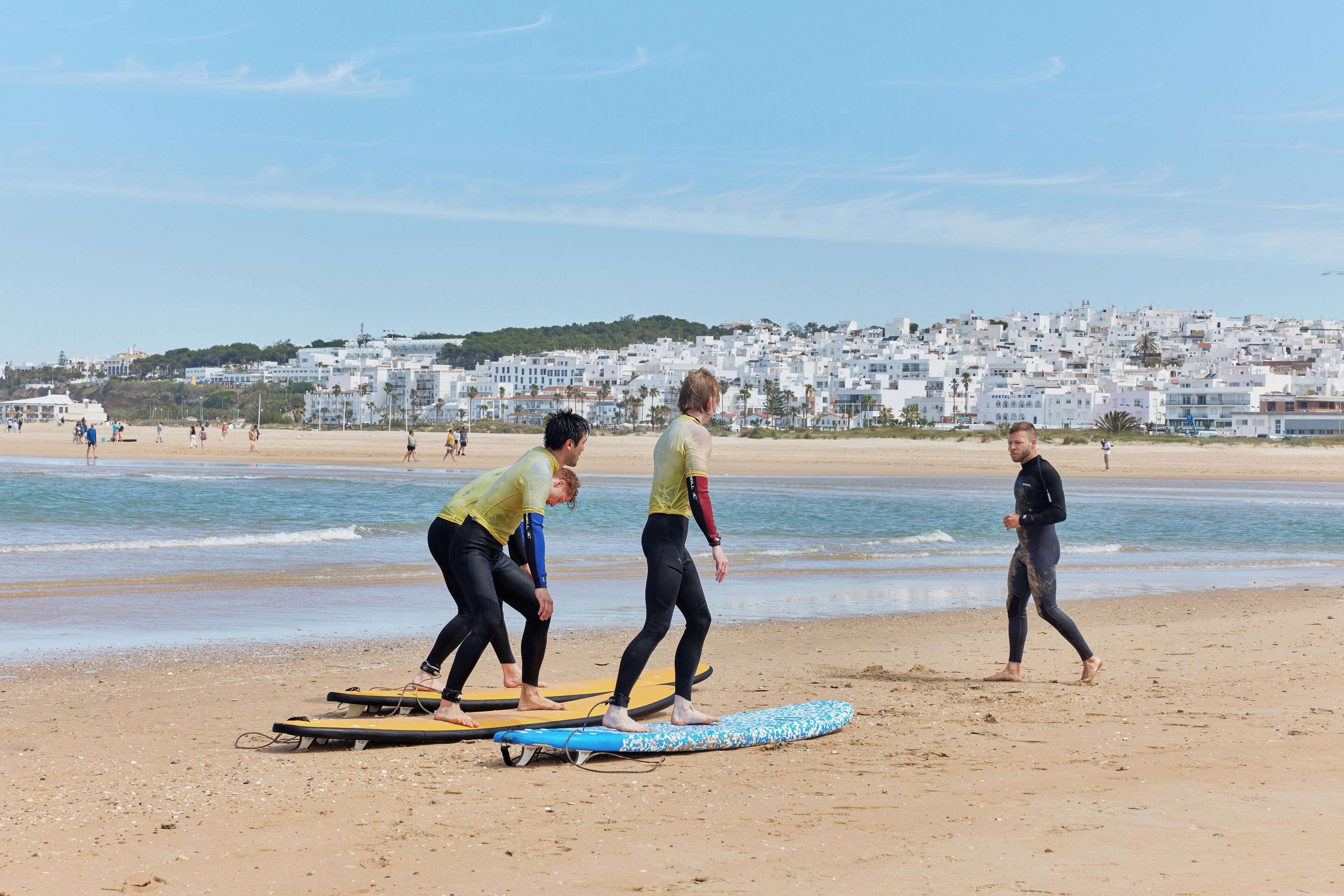 Een strand met mensen die surfboards klaarmaken, een heldere lucht en een verafgelegen stadsbeeld.