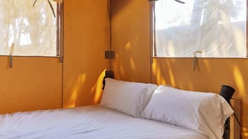 Un lit simple avec une tête de lit en bois, dans une pièce dotée de deux fenêtres, chacune étant pourvue d’un rideau.