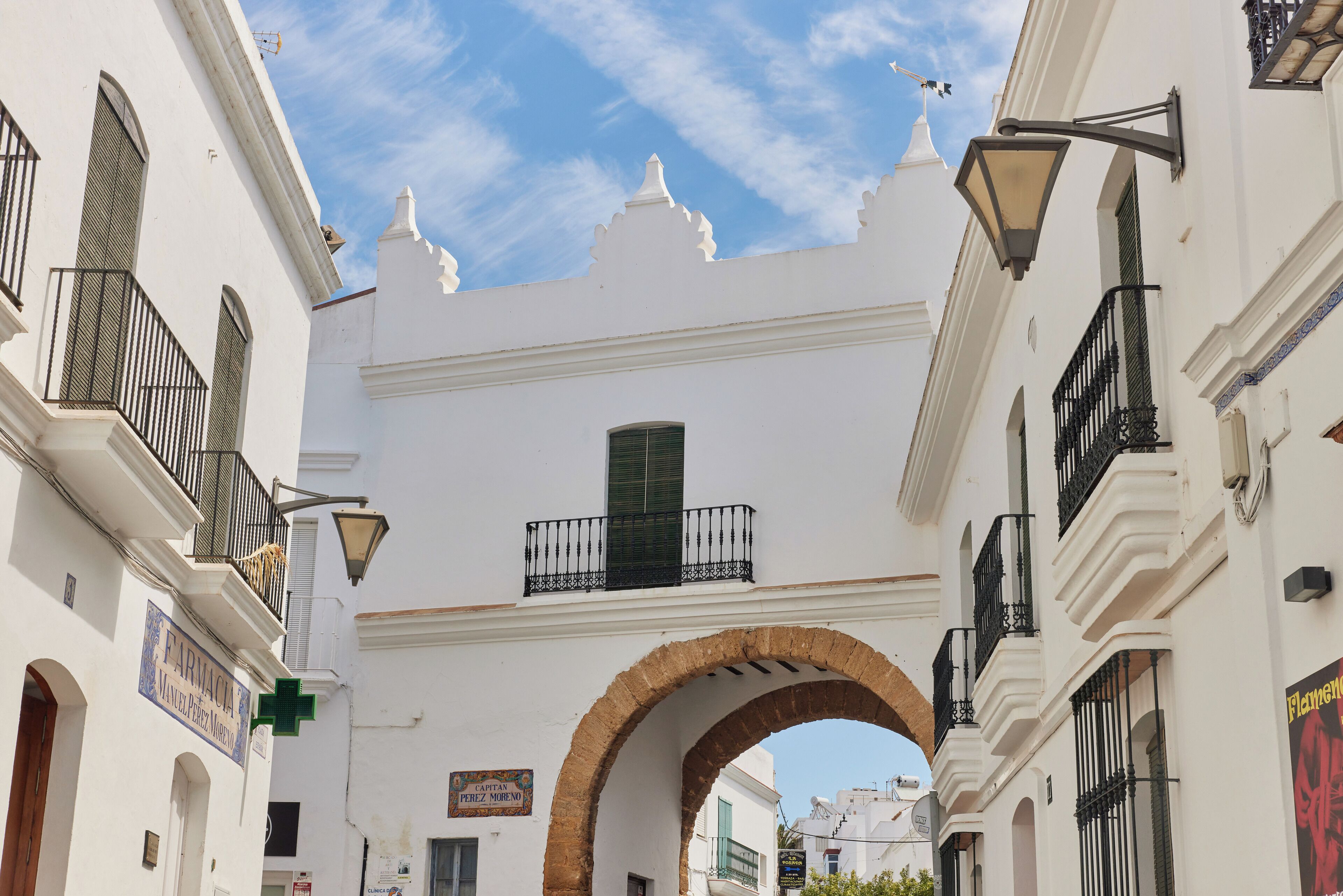 A narrow street with white buildings, a brick archway, and a sign for 'FARMACIA'.