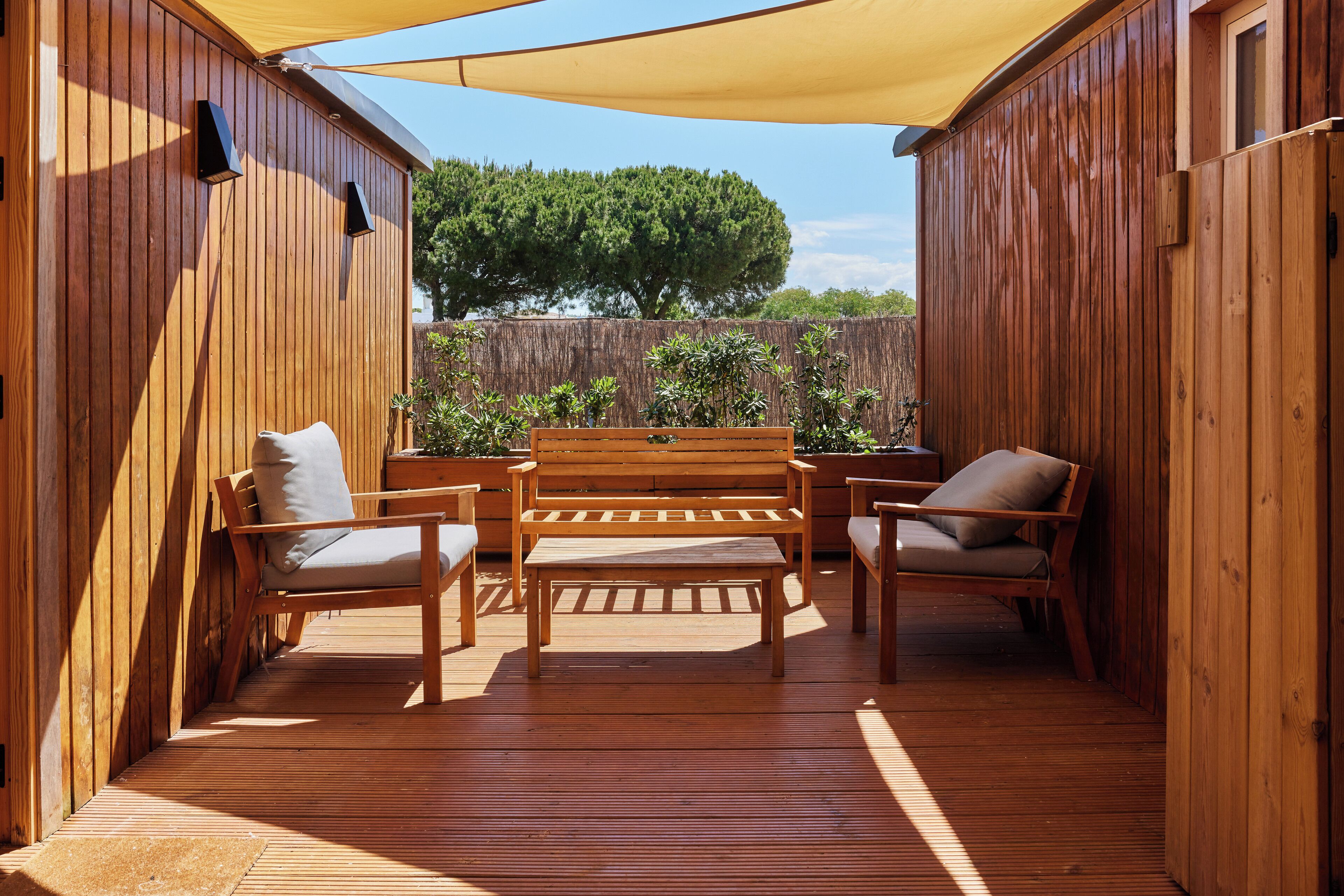 Una terraza de madera con un banco y dos sillas, rodeada de paredes de madera y un toldo grande.