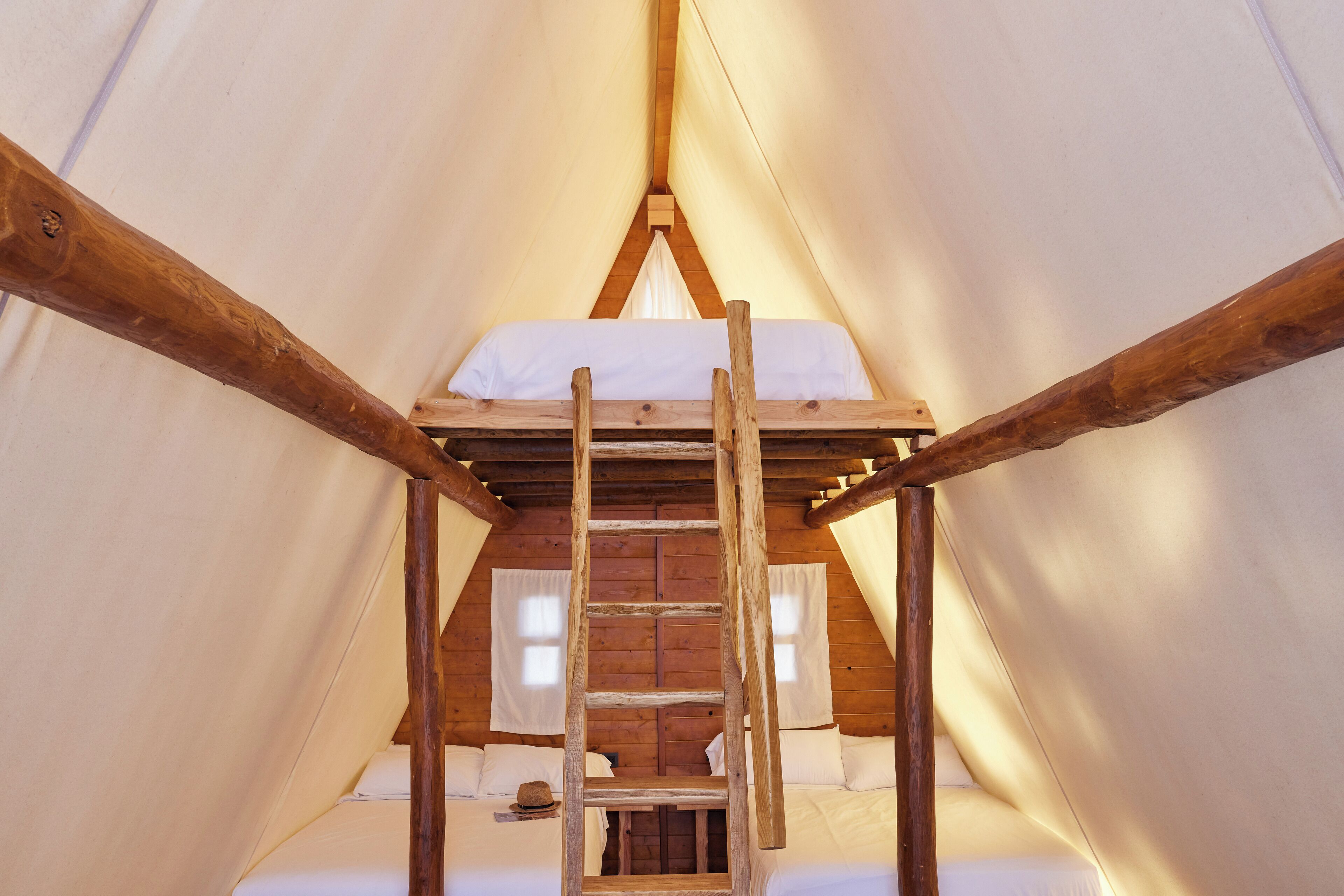 Une tente avec une échelle en bois menant à un lit en mezzanine, du linge de lit blanc et une petite fenêtre.
