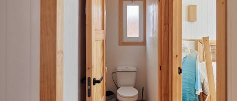 A narrow wooden hallway leading to a small bathroom with a toilet, a window, and a door.