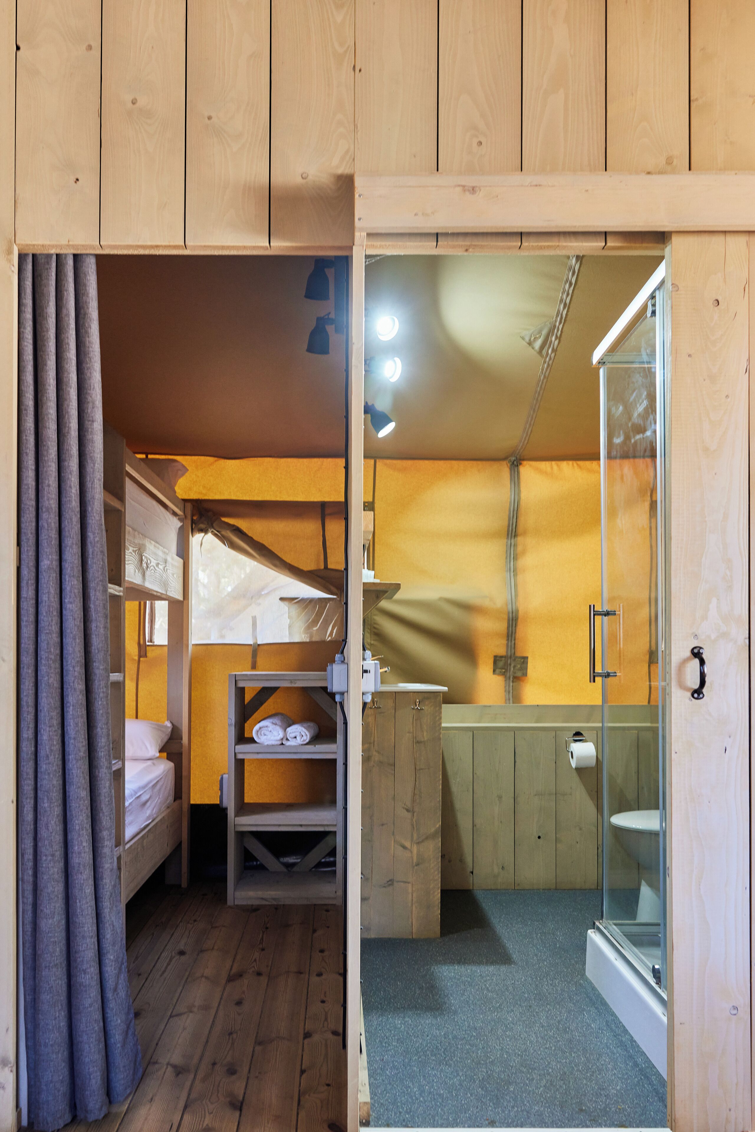 Une petite pièce comprenant un lit superposé, une petite étagère avec des serviettes, une armoire en bois et une cabine de douche en verre.