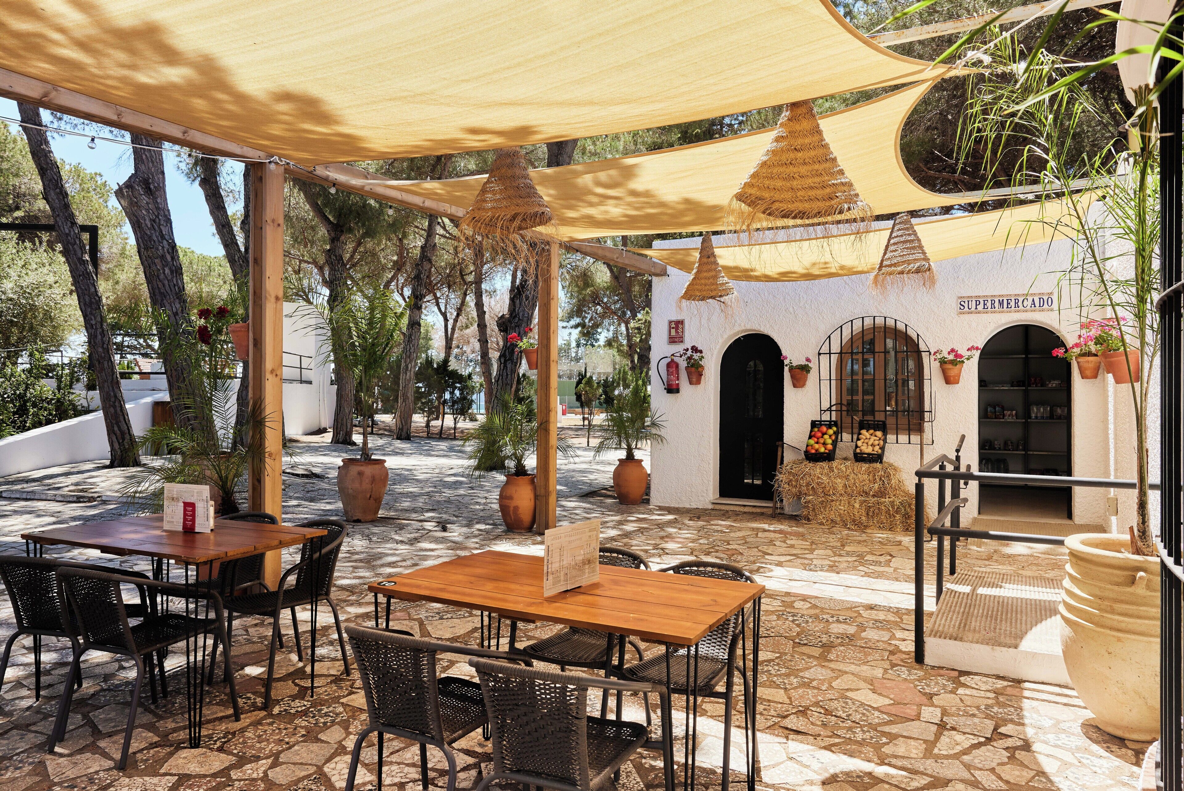 Une terrasse extérieure avec des tables et des chaises en bois, entourée de plantes en pot et d’un bâtiment blanc sur lequel on peut lire l’inscription « SUPERMERCADO ».