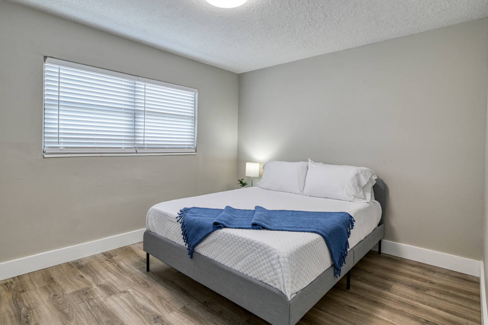 A bedroom with a bed, a window with blinds, a bedside lamp, and a wooden floor.