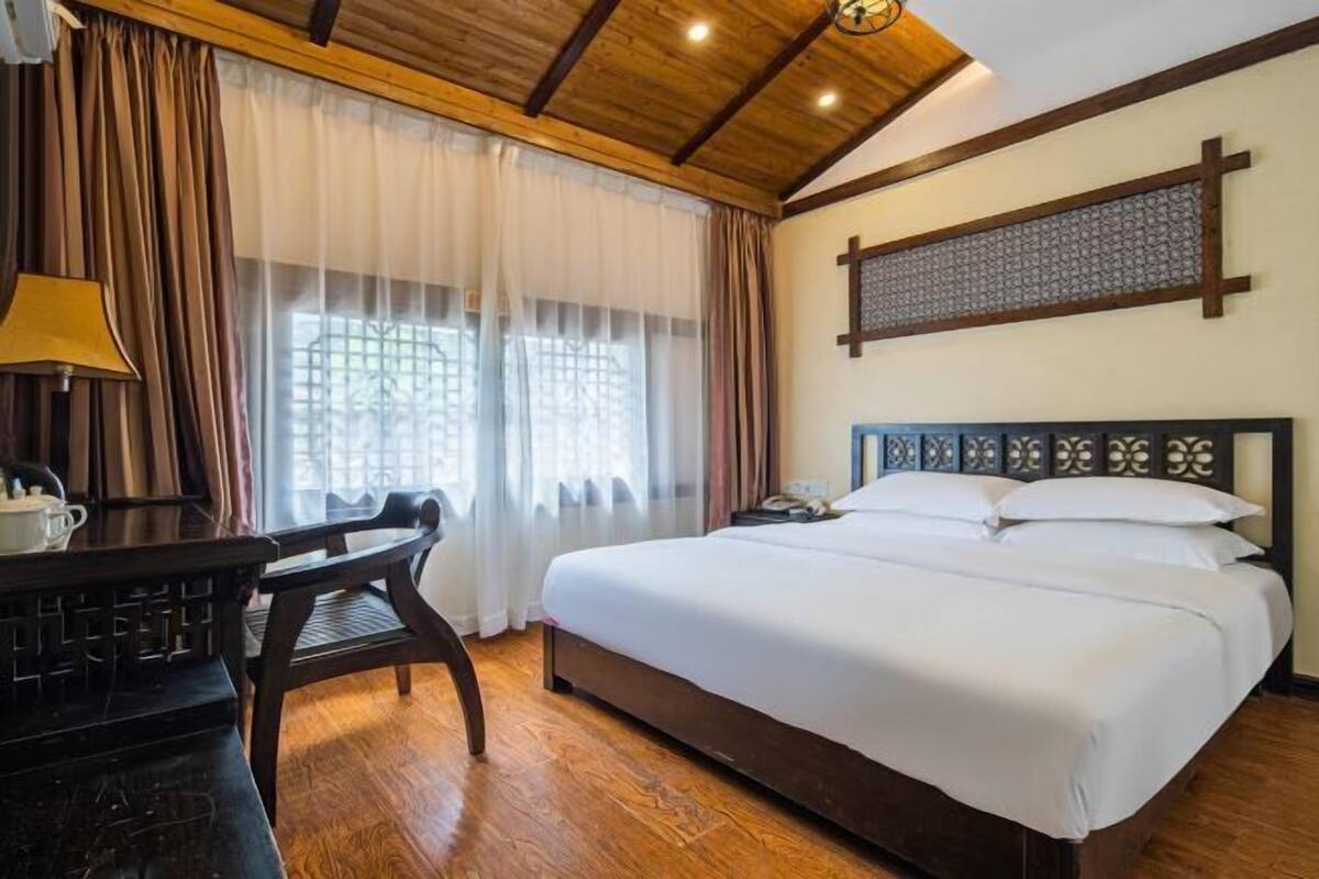 Une chambre d’hôtel avec un plafond en bois, un grand lit avec du linge de lit blanc, un bureau avec une lampe et une fenêtre avec des rideaux.