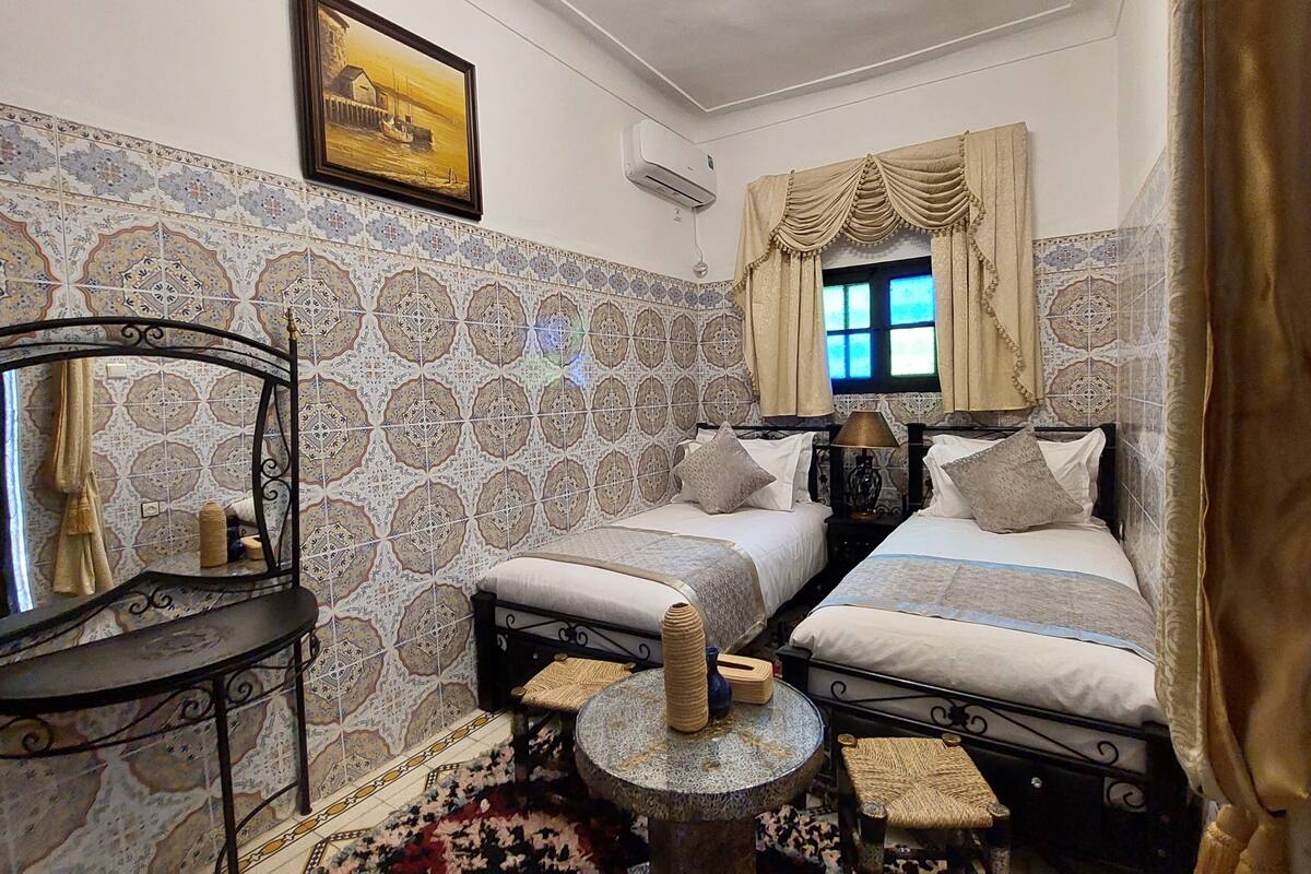 A room with two beds, a round table, a mirror, patterned walls, a window with curtains, and an air conditioning unit.