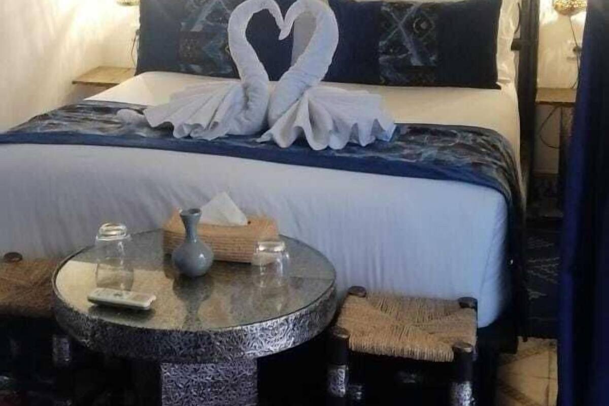 A neatly made bed with a heart-shaped pillow, a small round table with a vase and glasses, and a colorful rug.