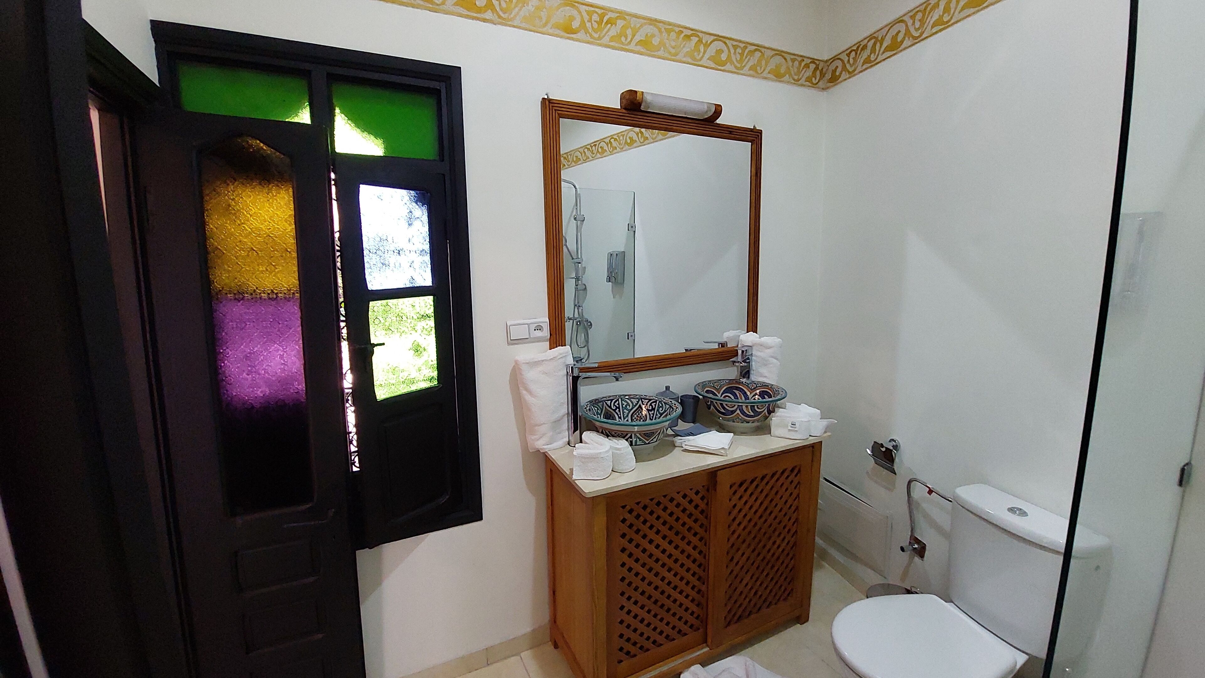 Une salle de bain comprenant des toilettes, un lavabo avec deux cuvettes décoratives, un miroir et une porte ornée d’un vitrail.