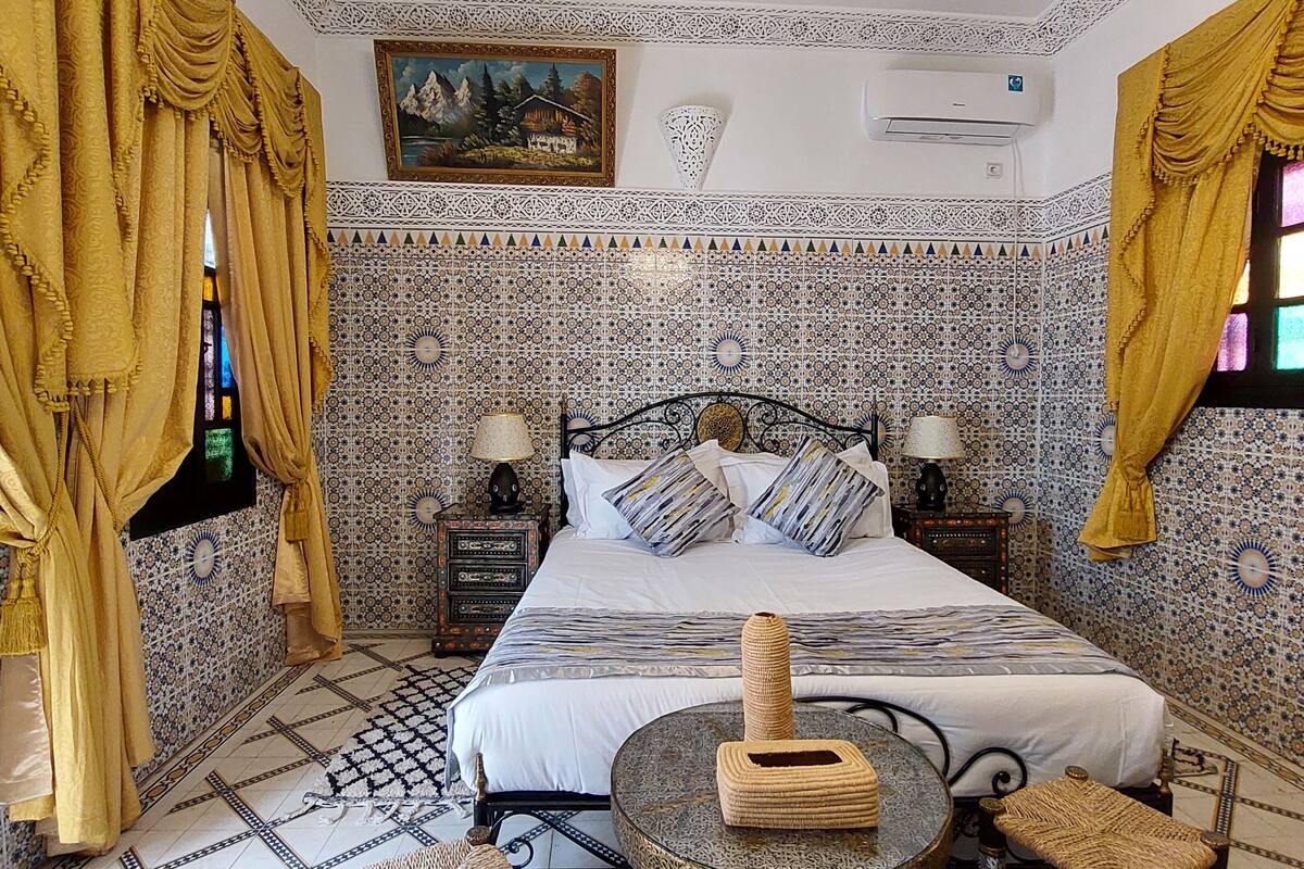 A room with a bed, two nightstands, a round table, and a window with curtains.
