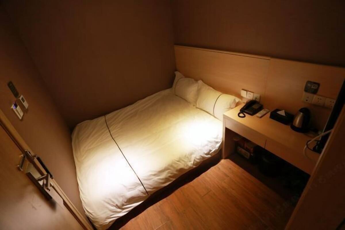 A small, single-bed room with a wooden floor, a bedside table with a phone and a lamp, and a wall-mounted power outlet.