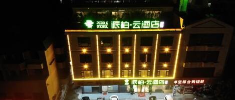 Entrada de hotel con un letrero, varios coches estacionados frente a ella y un edificio con ventanas iluminadas.