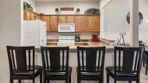 A modern kitchen with wooden cabinets, a white refrigerator, a microwave, and a coffee maker. There are four black chairs in front of the kitchen island. - Silver Dollar Hideaway in Stonebridge Community (Branson West)