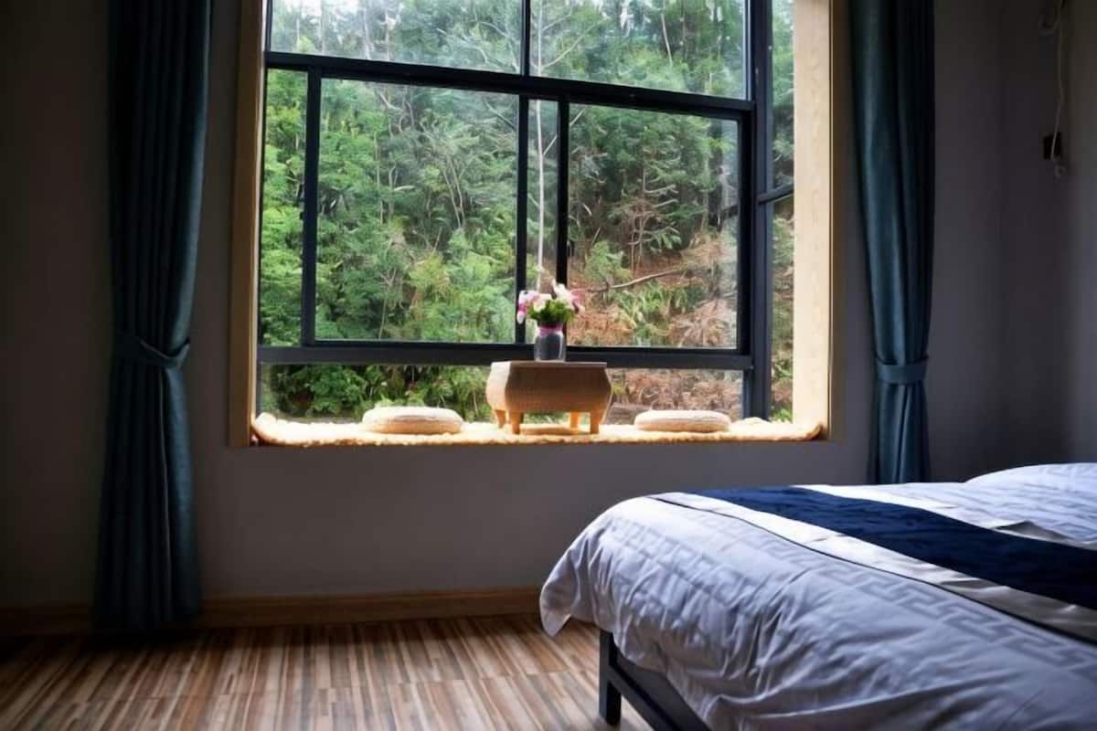 Ein Schlafzimmer mit großem Fenster, einem Bett und einem kleinen Hocker mit einer Vase.