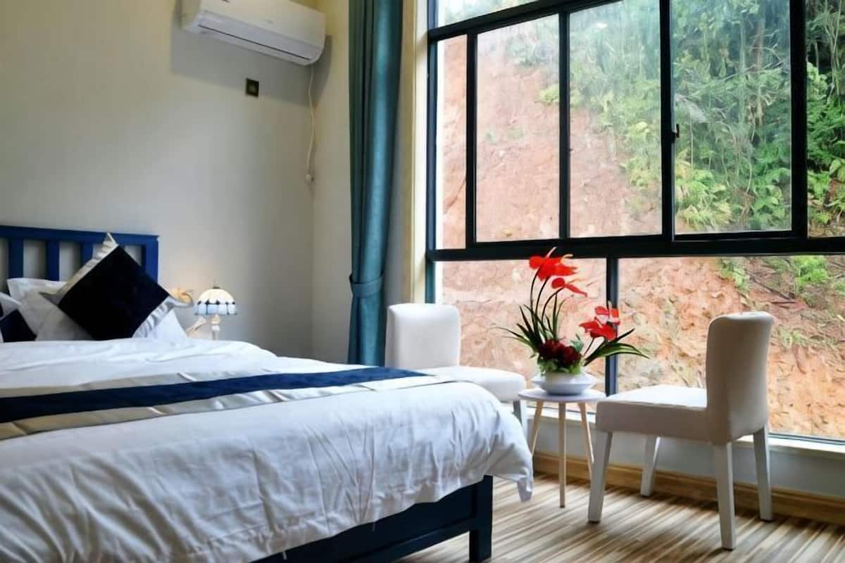 Ein Schlafzimmer mit einem Bett, einem kleinen Tisch mit Blumen in einer Vase, einem Sessel und einem großen Fenster mit Blick auf Bäume.