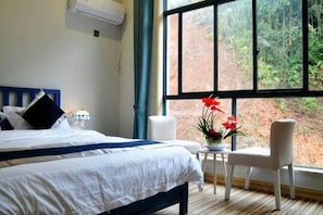A bedroom with a bed, a small table with a vase of flowers, a chair, and a large window with a view of trees. - Huashu Theme Inn (Wuyuan)
