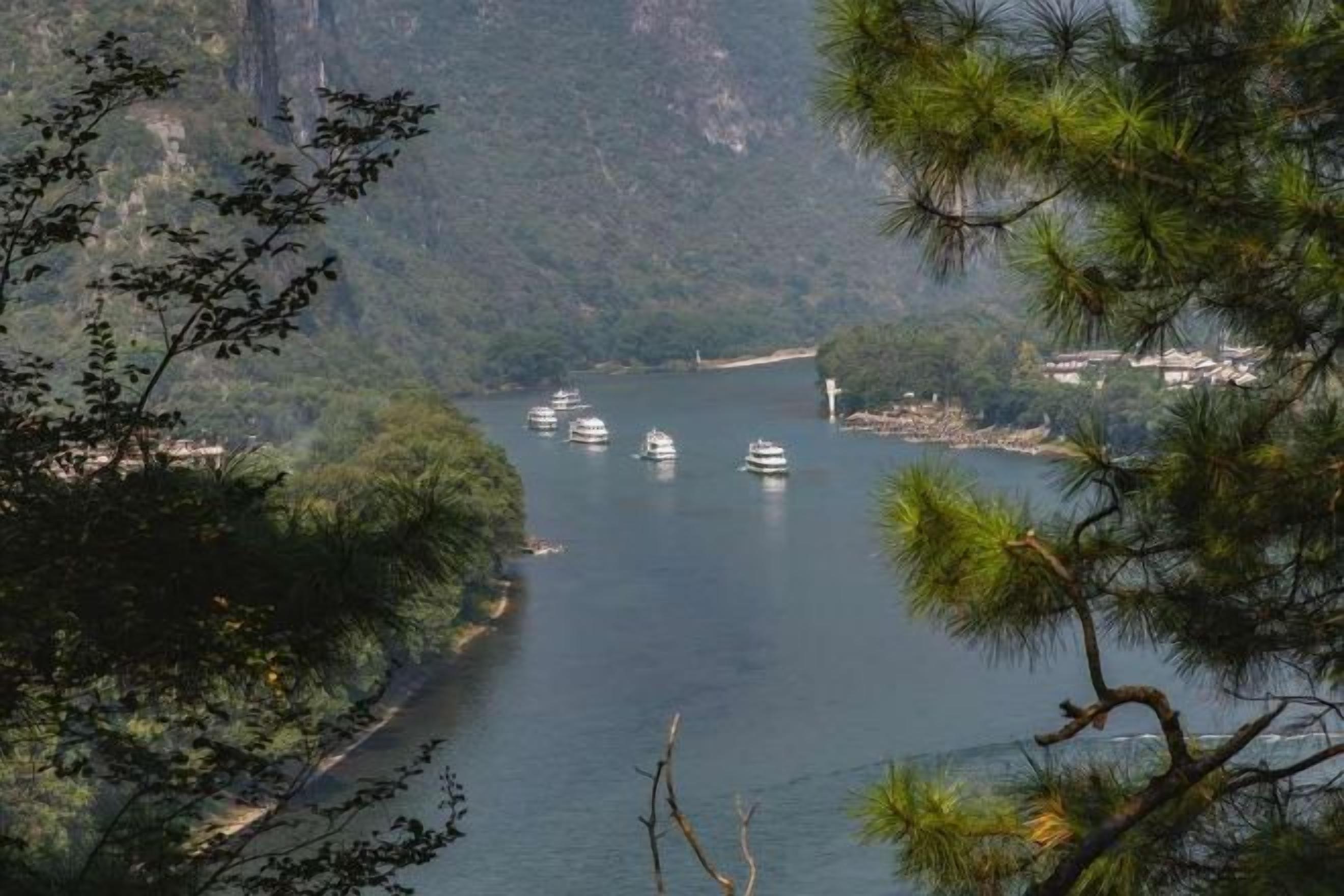 A river with boats, surrounded by forested hills and a distant settlement.