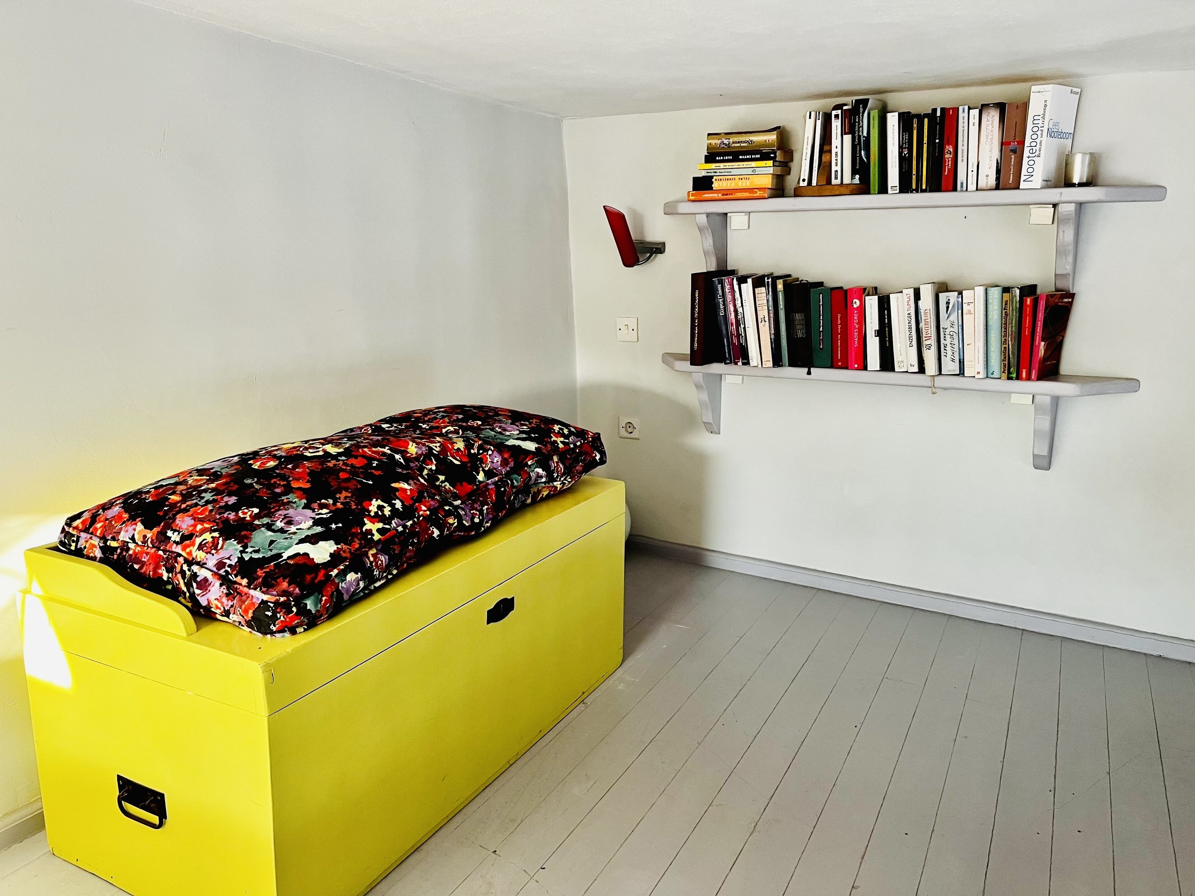 Une pièce avec un meuble de rangement jaune, un lit recouvert d’une housse à motifs floraux et deux étagères blanches remplies de livres.