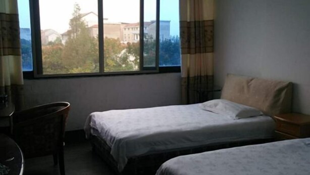 A room with two beds, a chair, a small table, and a window with a view of buildings and trees. - Kangfulai Hotel (Shengzhou)