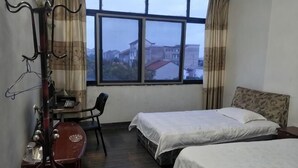 A hotel room with two beds, a desk, a chair, and a view of a building through the window. - Kangfulai Hotel (Shengzhou)
