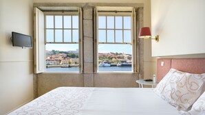 Habitación de hotel con cama, televisión, lámpara y vistas a la ciudad a través de dos ventanas.
