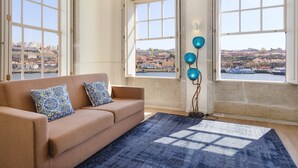 Una habitación con sofá color beige, cojines estampados, alfombra azul y vistas a un paisaje urbano a través de grandes ventanales.