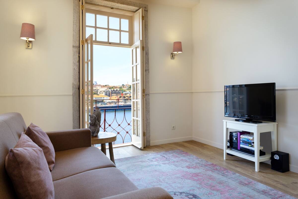 A living room with a sofa, a television on a stand, a patterned rug, and a view of the city through an open door.