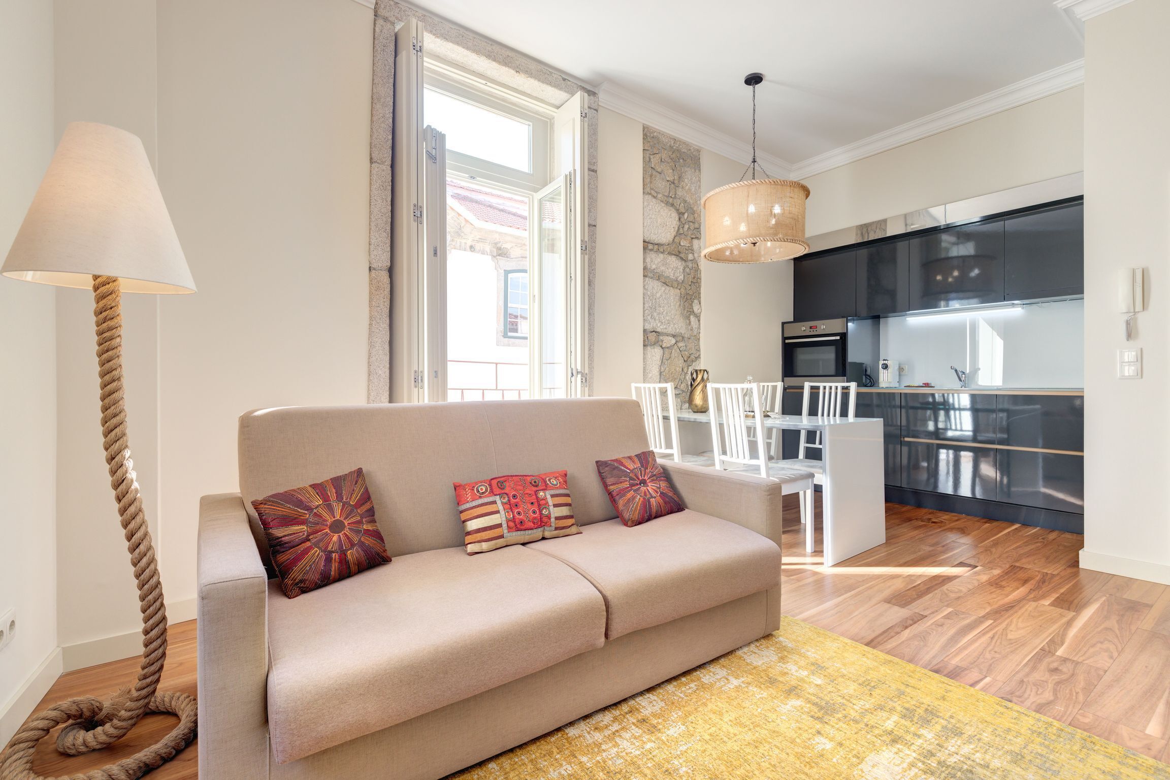 Ein Wohnzimmer mit einem beigefarbenen Sofa, zwei gemusterten Kissen, einem weißen Sessel, Holzboden und einer Küche im Hintergrund.