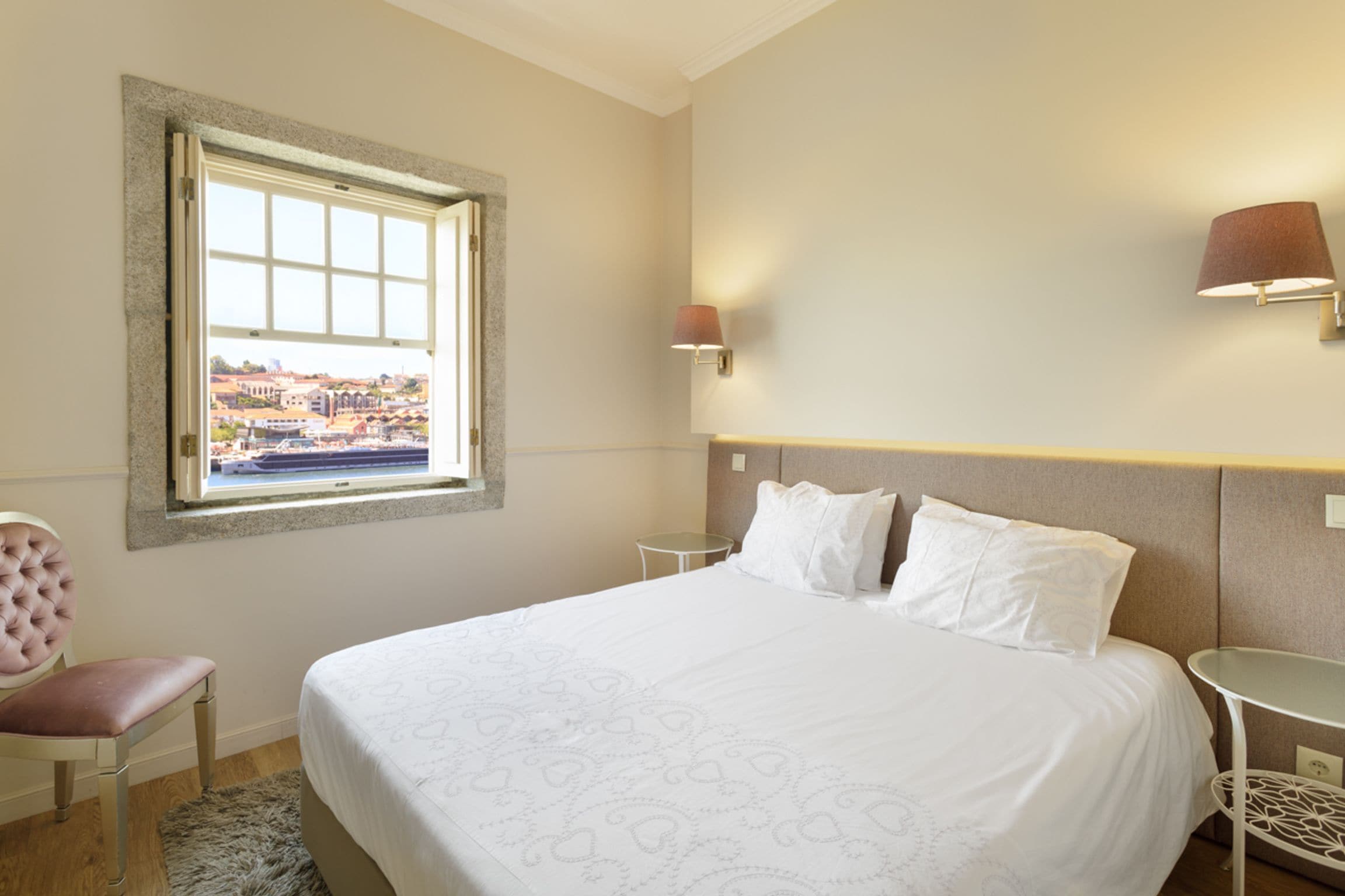 A hotel room with a large bed, a window with a view, a chair, and a small table.