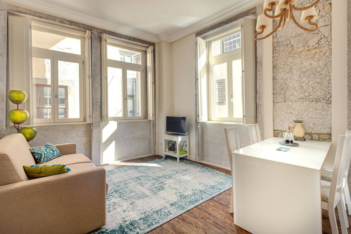 A room with a sofa, a small TV stand, a dining table, and a large window.